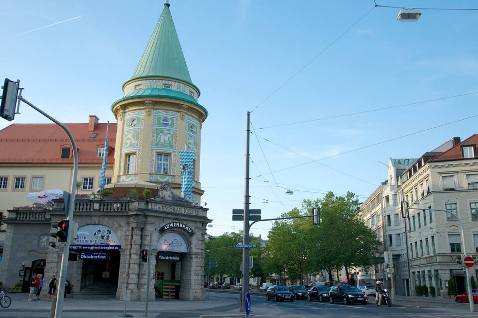 Una torre histórica con aguja verde, una calle con coches aparcados y un edificio con un cartel de Oktoberfest.