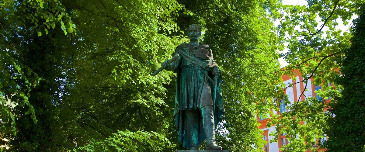 A statue of a historical figure surrounded by lush green trees and a colorful building in the background.