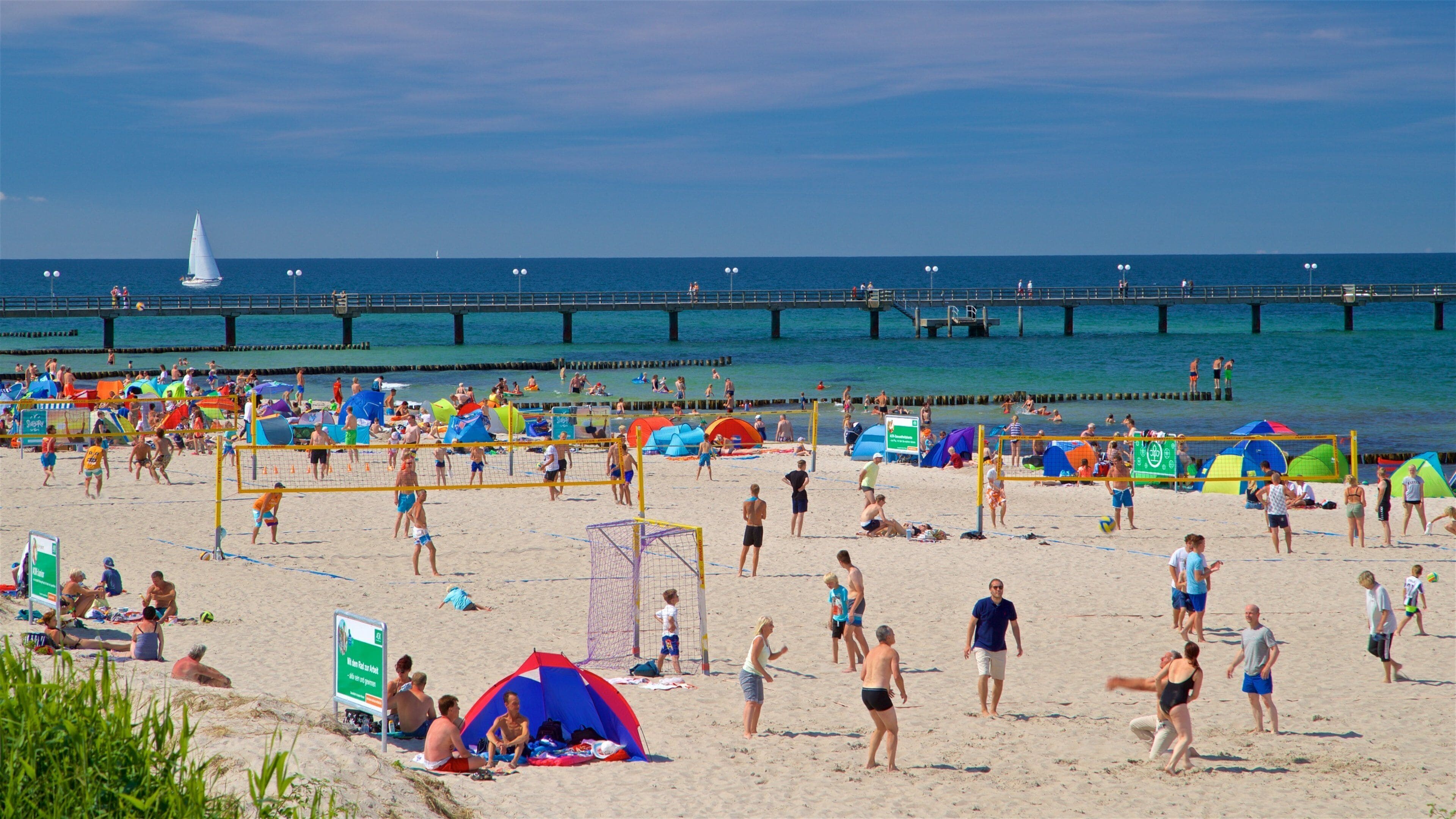 A crowded beach with people playing volleyball, tents, and a pier in the background.