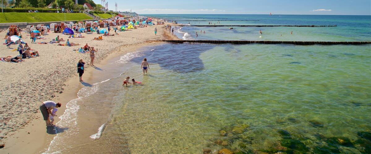 A crowded beach with clear shallow waters, people sunbathing, and a distant sailboat.