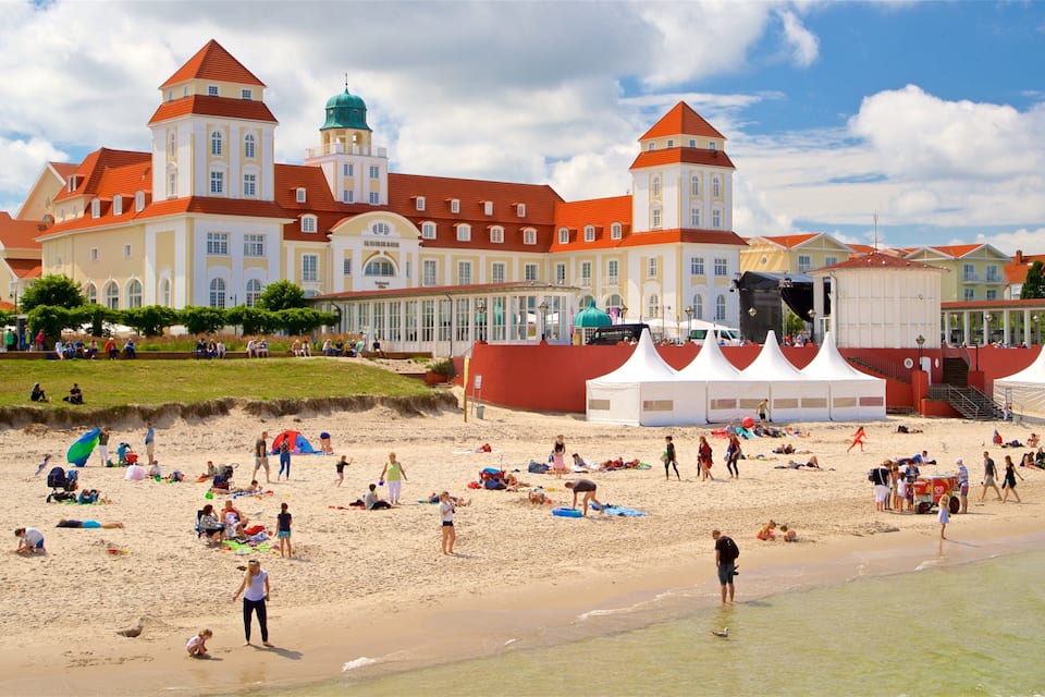 Een strand met mensen die zonnebaden en spelen, een groot gebouw met rode daken, en tenten die zijn opgezet voor een evenement.