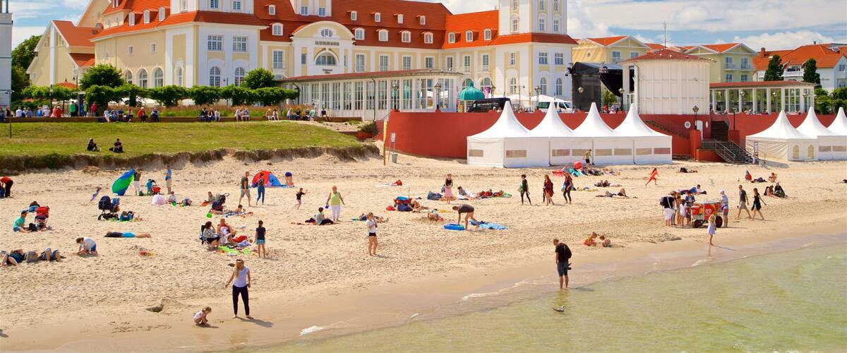 A beach with people sunbathing and playing, a large building with red roofs, and tents set up for an event.