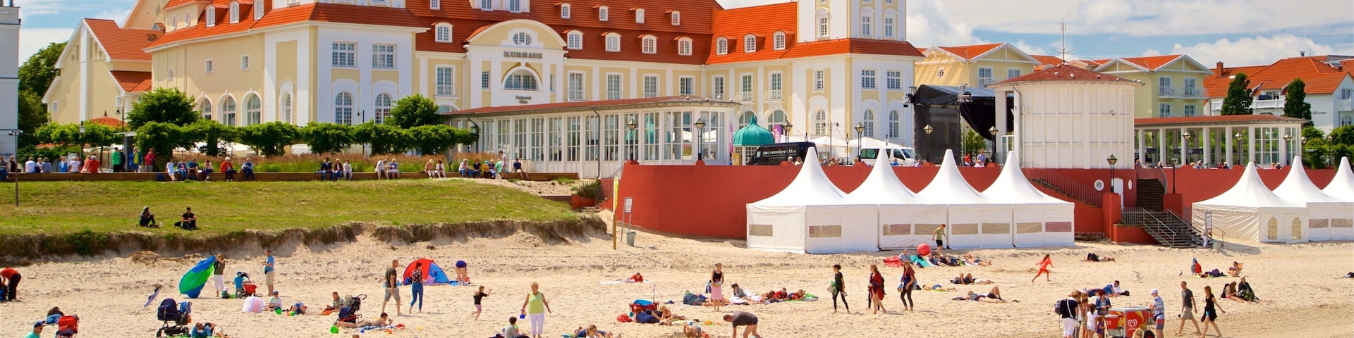 A beach with people sunbathing and playing, a large building with red roofs, and tents set up for an event.