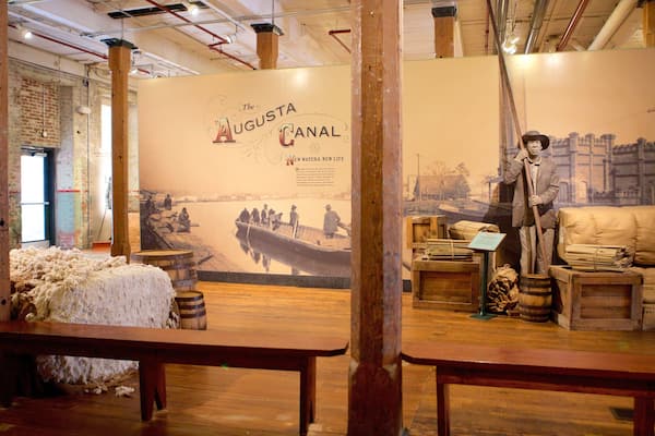 Une exposition historique comprenant une table en bois, une couverture en laine, des barils et une statue représentant un homme tenant une pelle.
