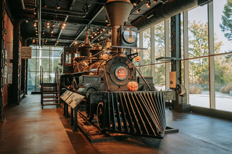 A vintage steam locomotive in a museum with large windows and a brick wall.
