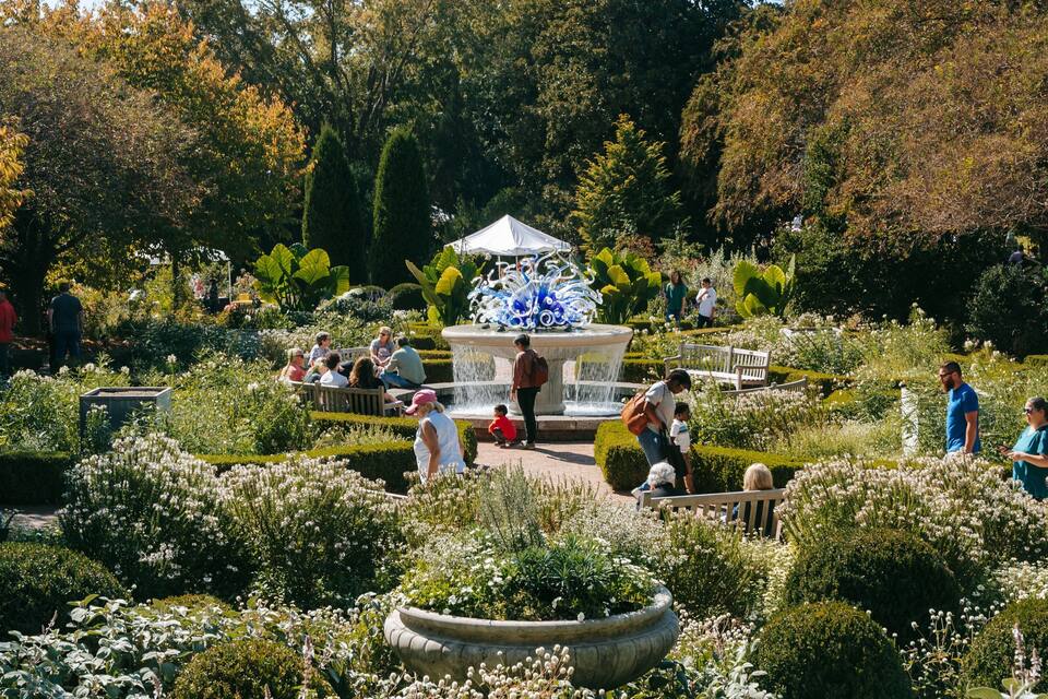 A garden with a fountain, benches, and people walking around.