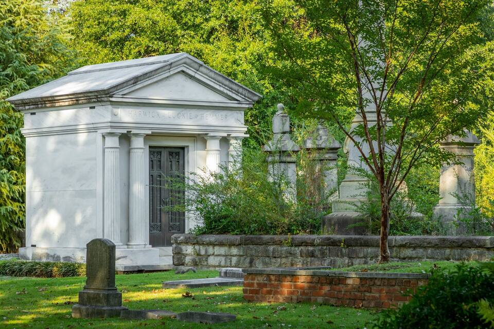 Ein weißes Marmormausoleum mit der Inschrift „Harwick Lucke Keyner“ in einem von Bäumen und Grünpflanzen umgebenen Friedhof.