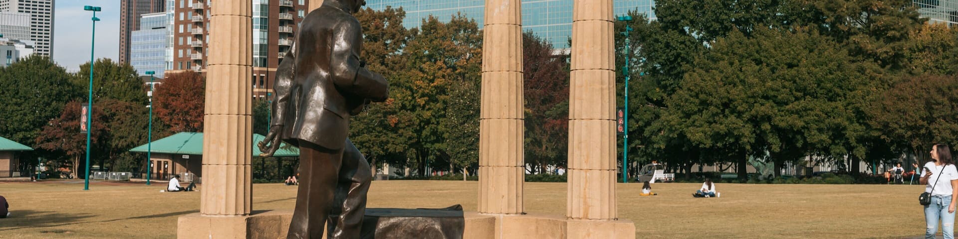 A bronze statue of a man in a suit, a metal structure with Olympic rings, and modern buildings in the background.