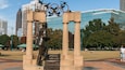 Una estatua de bronce de un hombre con traje, una estructura metálica con aros olímpicos y edificios modernos al fondo.