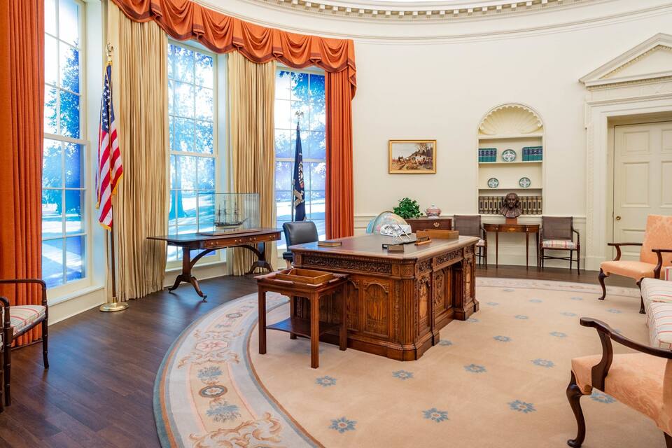 A formal room with a large wooden desk, two flags, and a large window with curtains.
