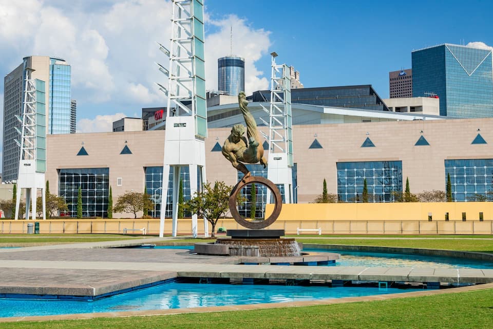 A sculpture of a man performing a handstand on a ring, a fountain, and a modern building with large windows and a yellow wall.