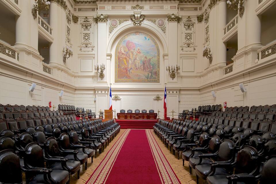 A grand hall with rows of chairs, a red carpet, and a large mural on the wall.