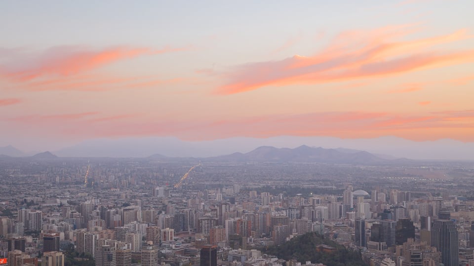 Paisagem urbana com céu ao pôr do sol, edifícios densos e montanhas distantes.