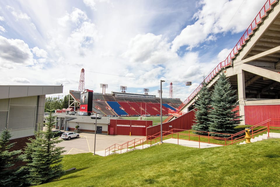 A stadium with red seating, a parking lot, and a grassy area with trees.