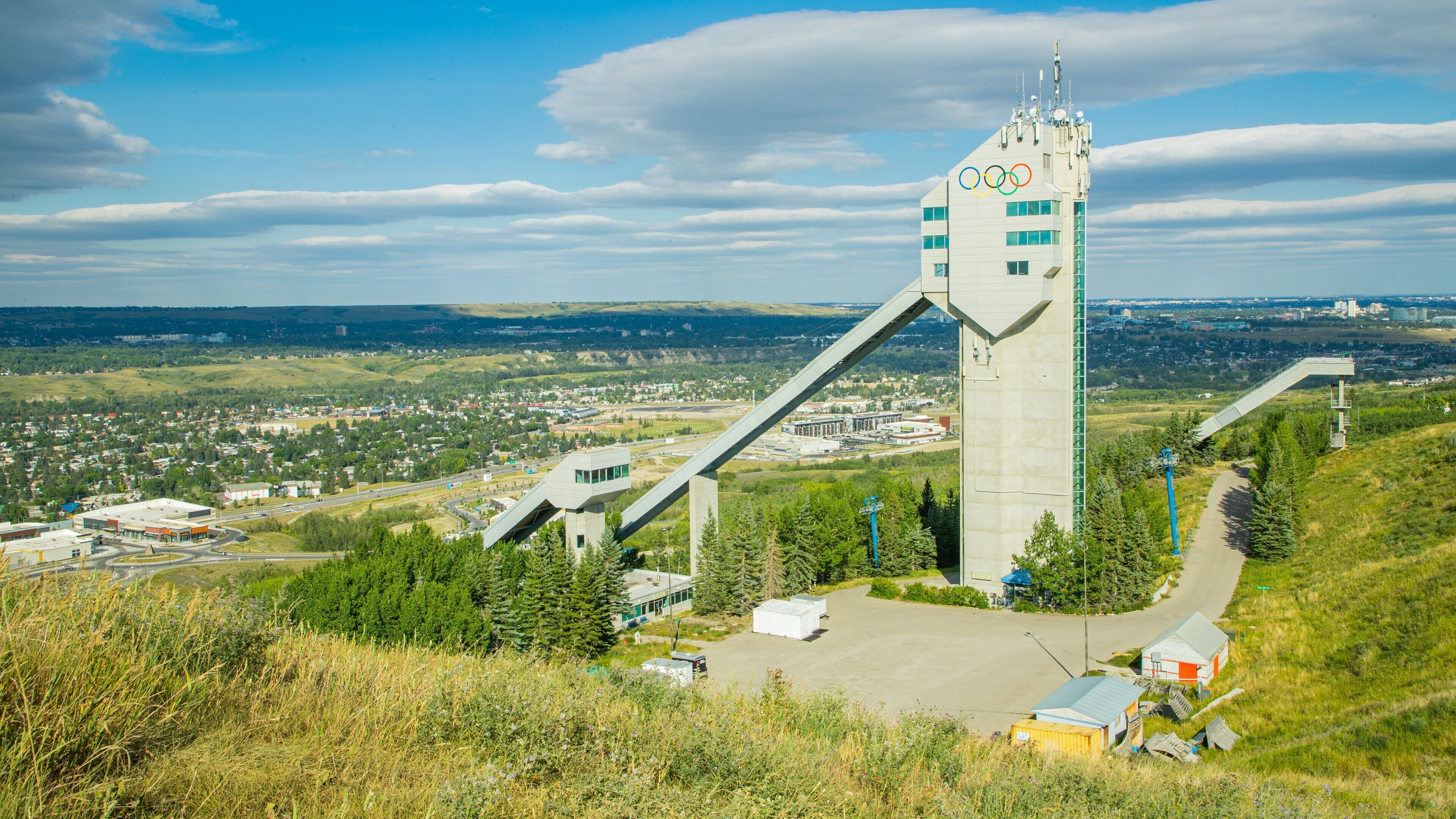 Une haute structure en béton surmontée des anneaux olympiques, entourée de verdure et avec une ville en arrière-plan.