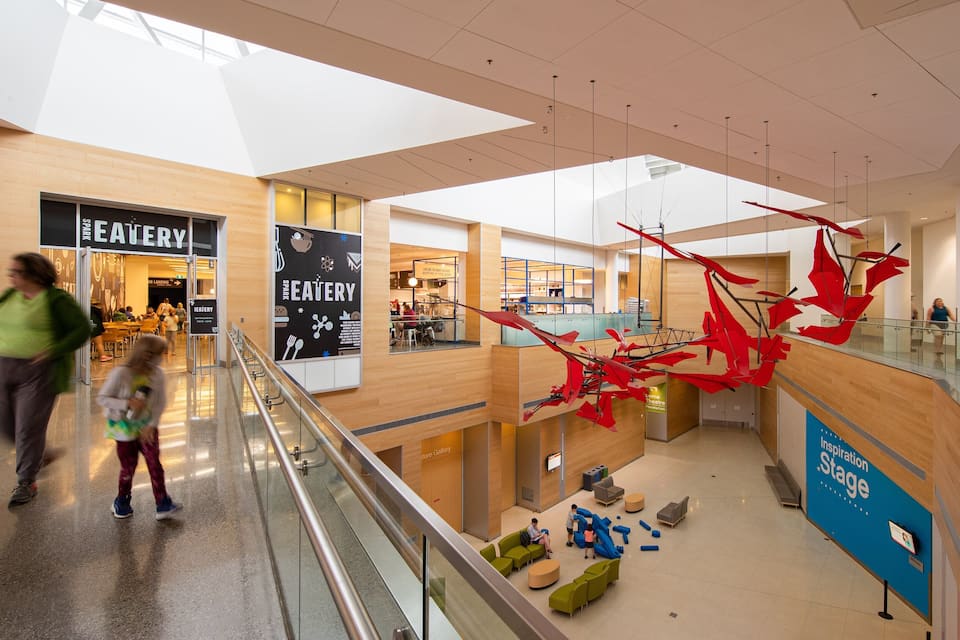 A shopping mall interior with a food court, a glass railing, and a red sculpture.