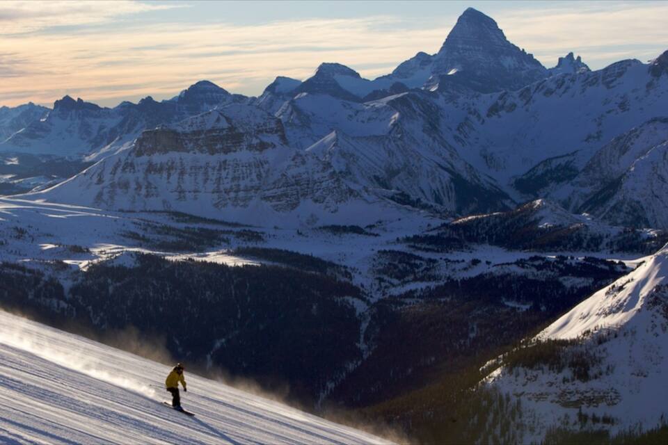 A snow-covered mountain range with a prominent peak in the background and a skier on a slope.