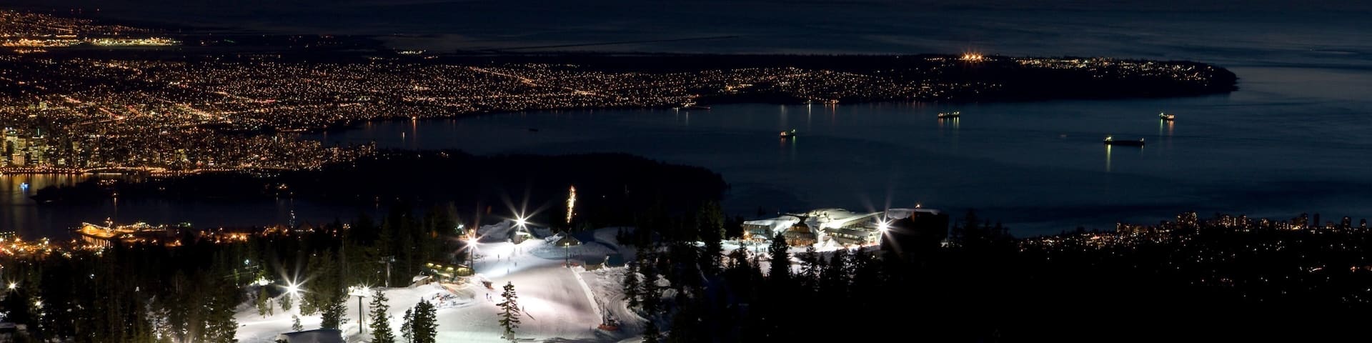 A snow-covered ski resort at dusk with illuminated ski lifts and a distant cityscape.