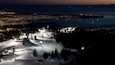 A snow-covered ski resort at dusk with illuminated ski lifts and a distant cityscape.