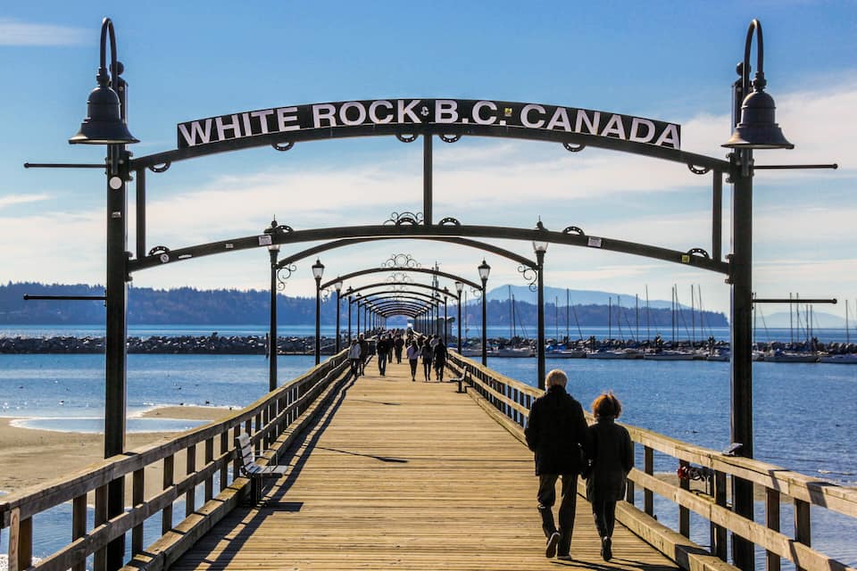 A wooden pier with a sign reading 'WHITE ROCK B.C. CANADA' leading to a body of water.
