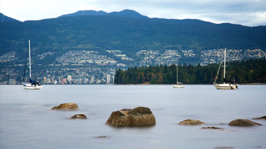 A calm lake with three sailboats, surrounded by mountains and a cityscape in the background.