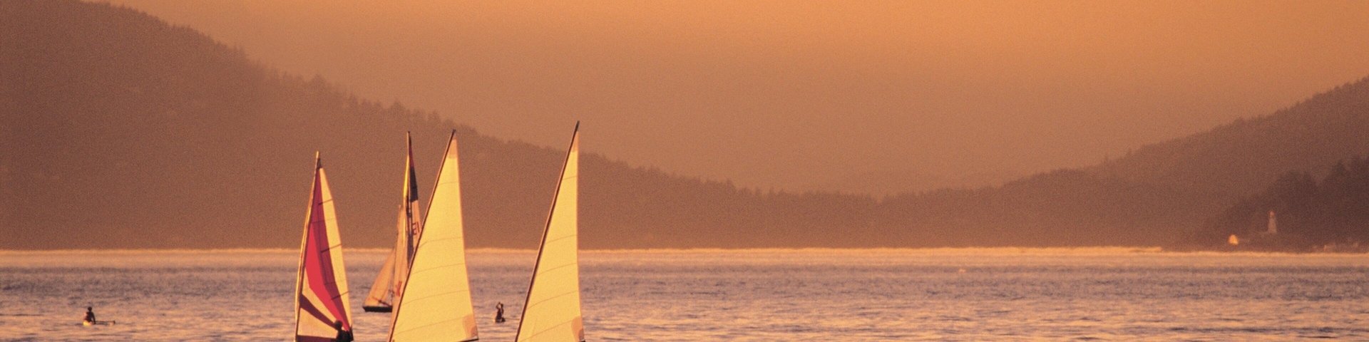 A calm lake with sailboats and kayaks, surrounded by mountains and a lighthouse in the distance.