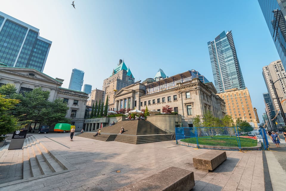 Uma praça urbana com um edifício histórico, arranha-céus modernos e céu claro.