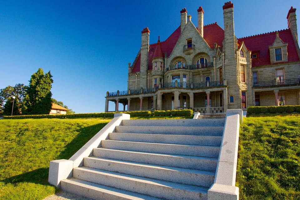 A grand, historic building with a red roof and stone architecture, surrounded by a well-manicured lawn and a set of stone steps leading up to the entrance.
