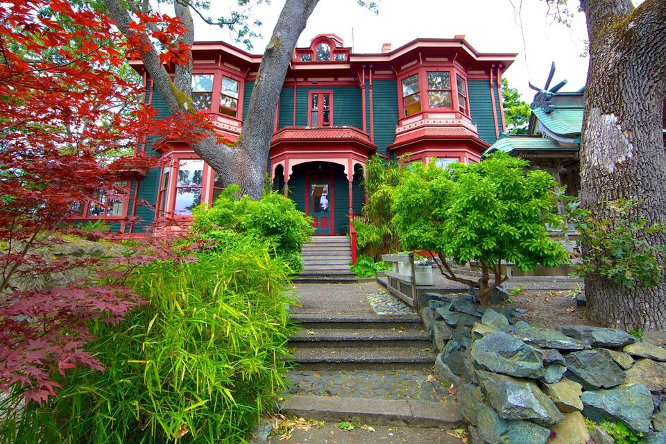 A Victorian-style house with red trim and green shutters, surrounded by lush foliage and stone steps leading to the entrance.