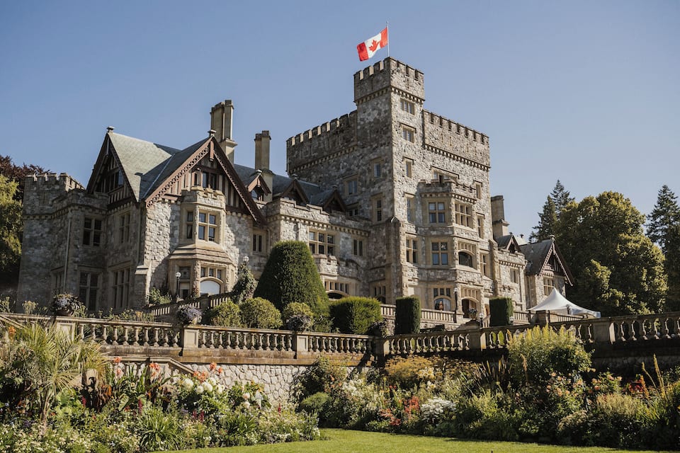 Um grande castelo de pedra com a bandeira do Canadá, rodeado por um jardim bem cuidado e uma ponte de pedra.
