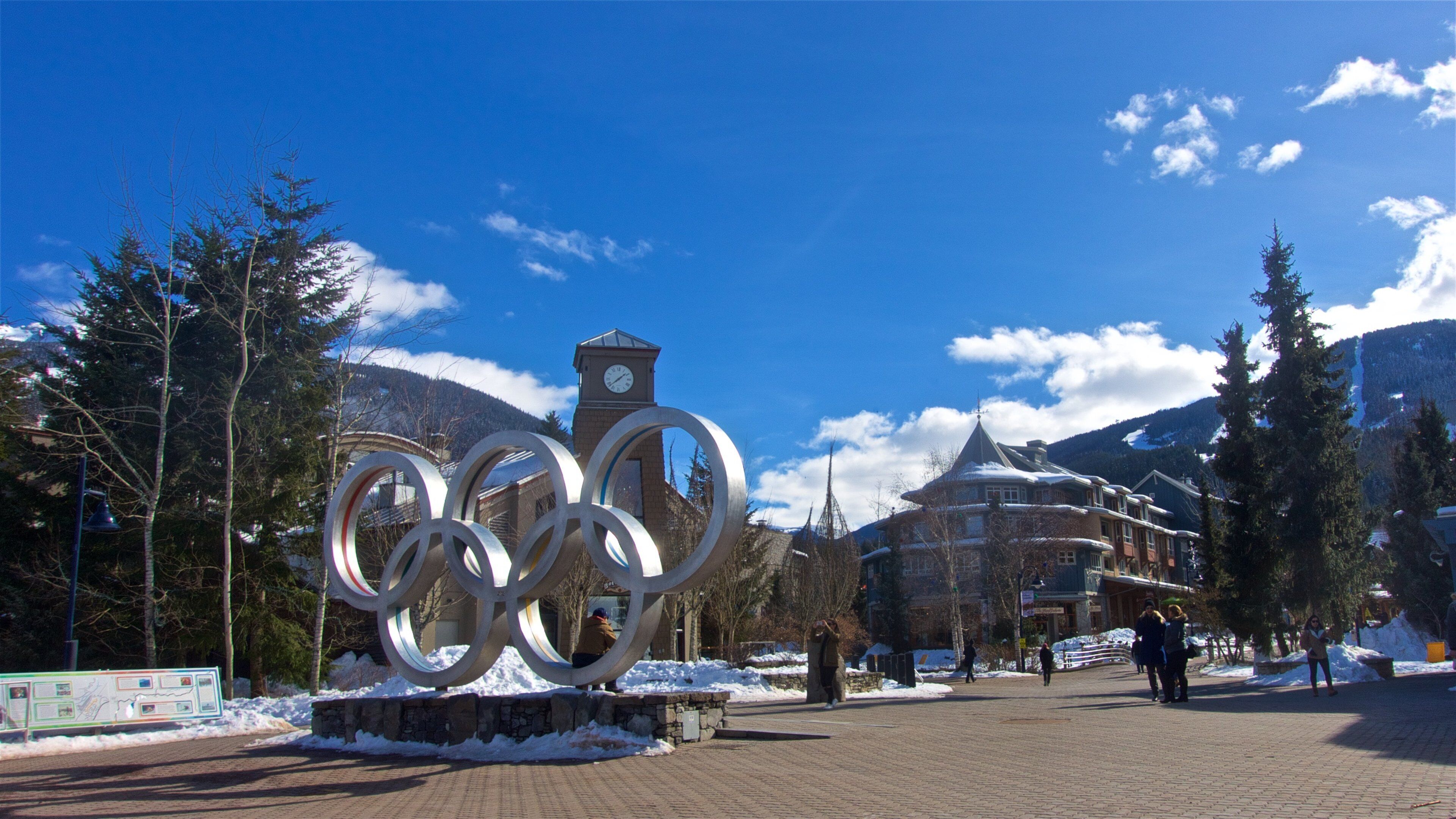 A snow-covered plaza with Olympic rings sculpture, a clock tower, and a mountain backdrop.