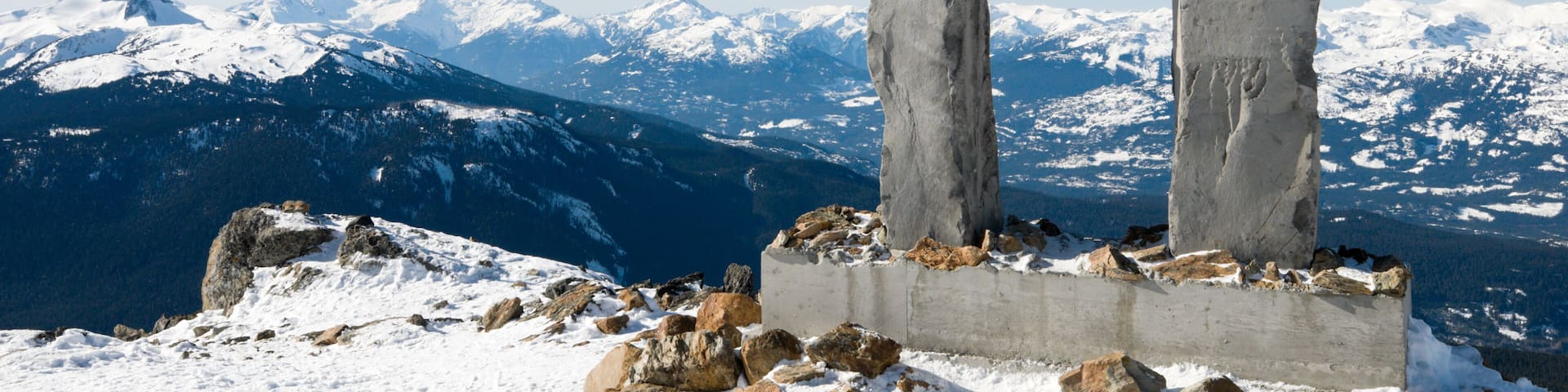 Un totém en la cima nevada de una montaña, con montañas cubiertas de nieve al fondo.