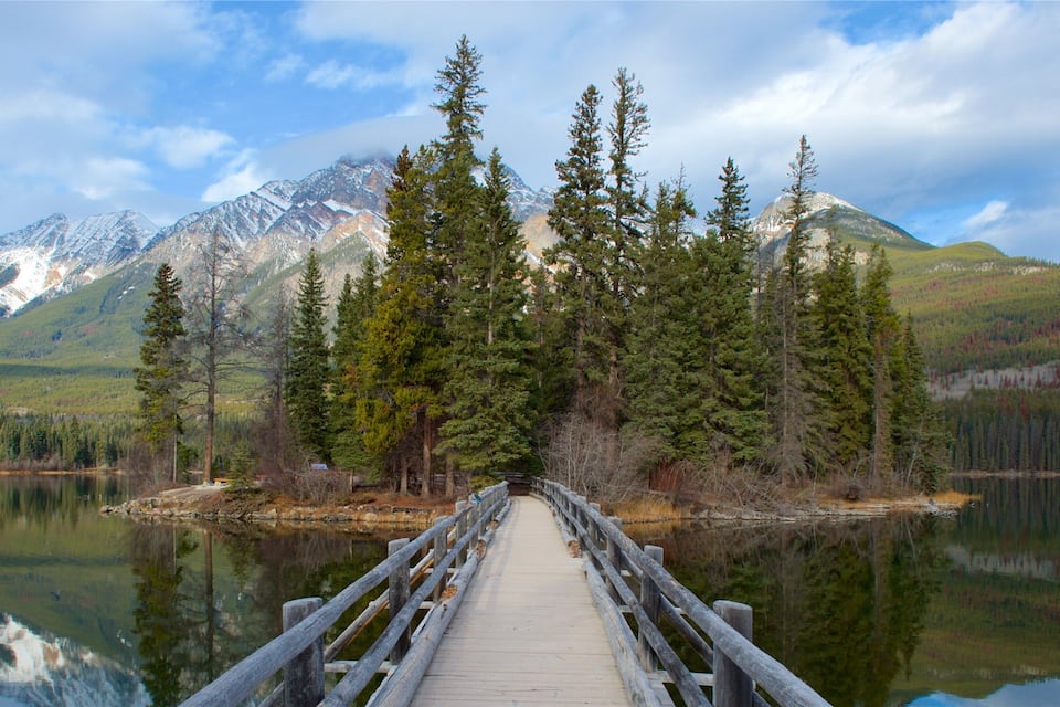 A wooden bridge over a calm lake with a forest and snow-capped mountains in the background.