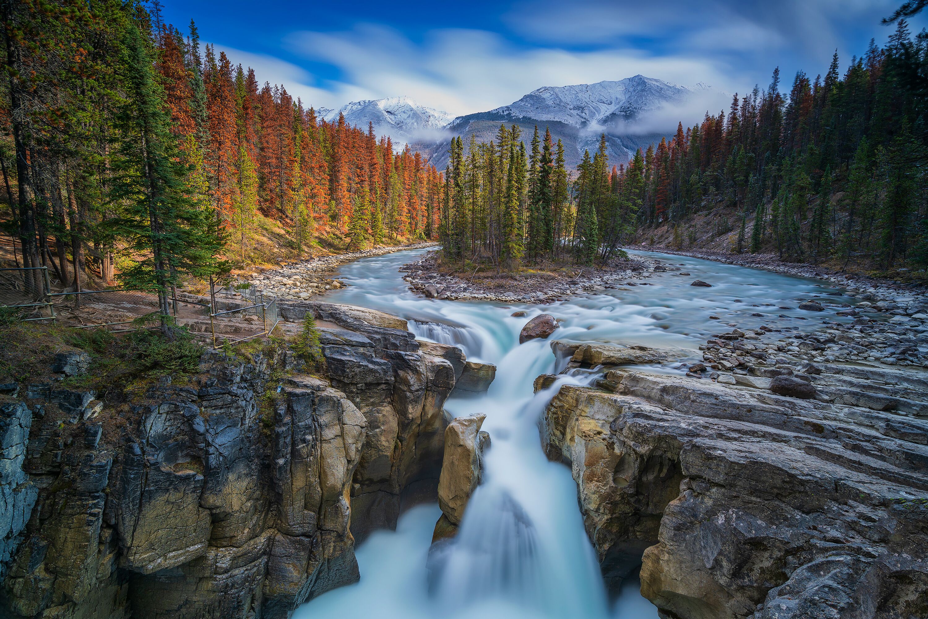 Ein Fluss fließt durch eine felsige Schlucht, umgeben von herbstlich gefärbten Bäumen und schneebedeckten Bergen im Hintergrund.