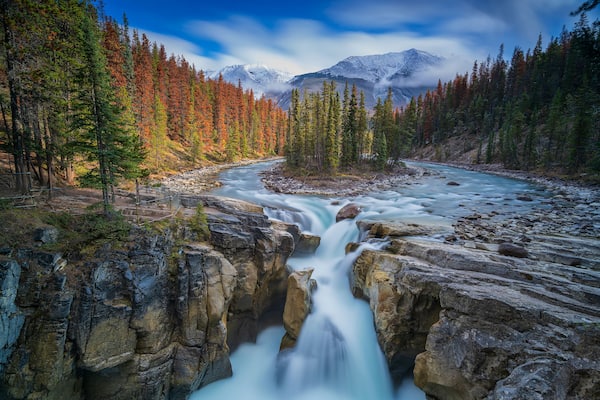 Ein Fluss fließt durch eine felsige Schlucht, umgeben von herbstlich gefärbten Bäumen und schneebedeckten Bergen im Hintergrund.