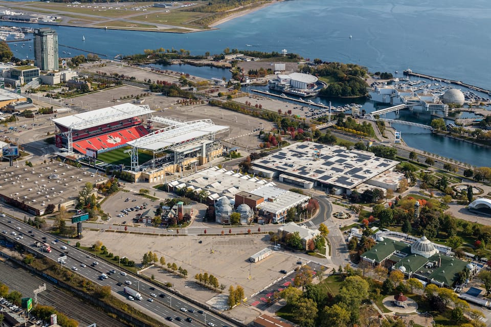 Vista aérea de una ciudad costera con un estadio grande, estacionamientos y un puerto deportivo con barcos.