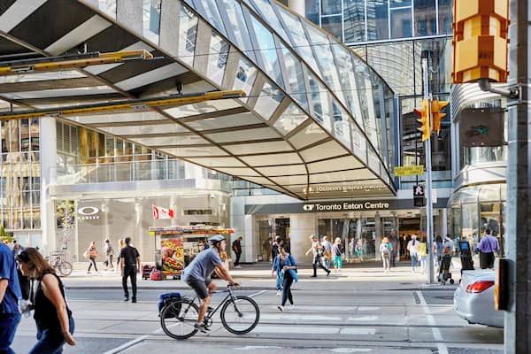 Une rue animée en milieu urbain, avec une passerelle piétonne couverte de verre, le centre commercial Toronto Eaton Centre, et un cycliste qui traverse.