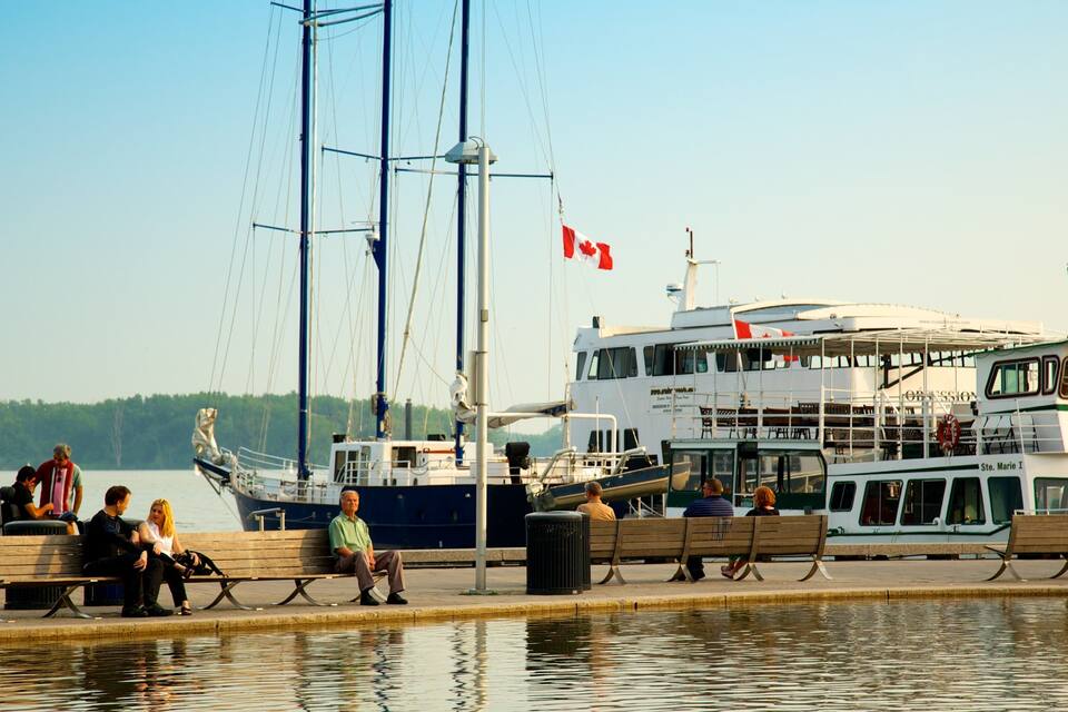 A waterfront with a large ferry named Ste. Marie I, people sitting on benches, and a sailboat in the background.