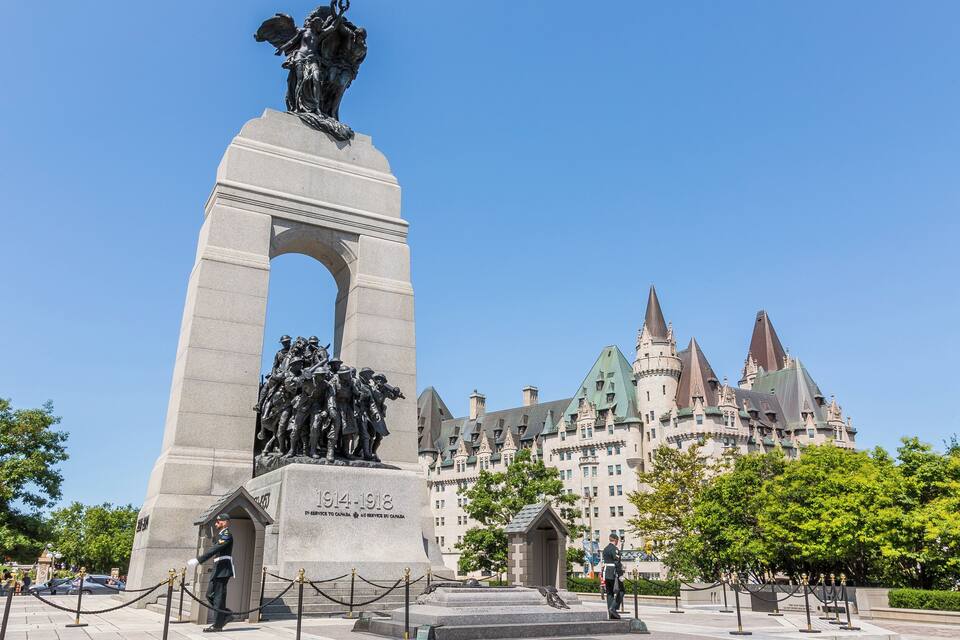 A monument with a statue of a soldier on top, surrounded by a fence and a building with multiple towers in the background.