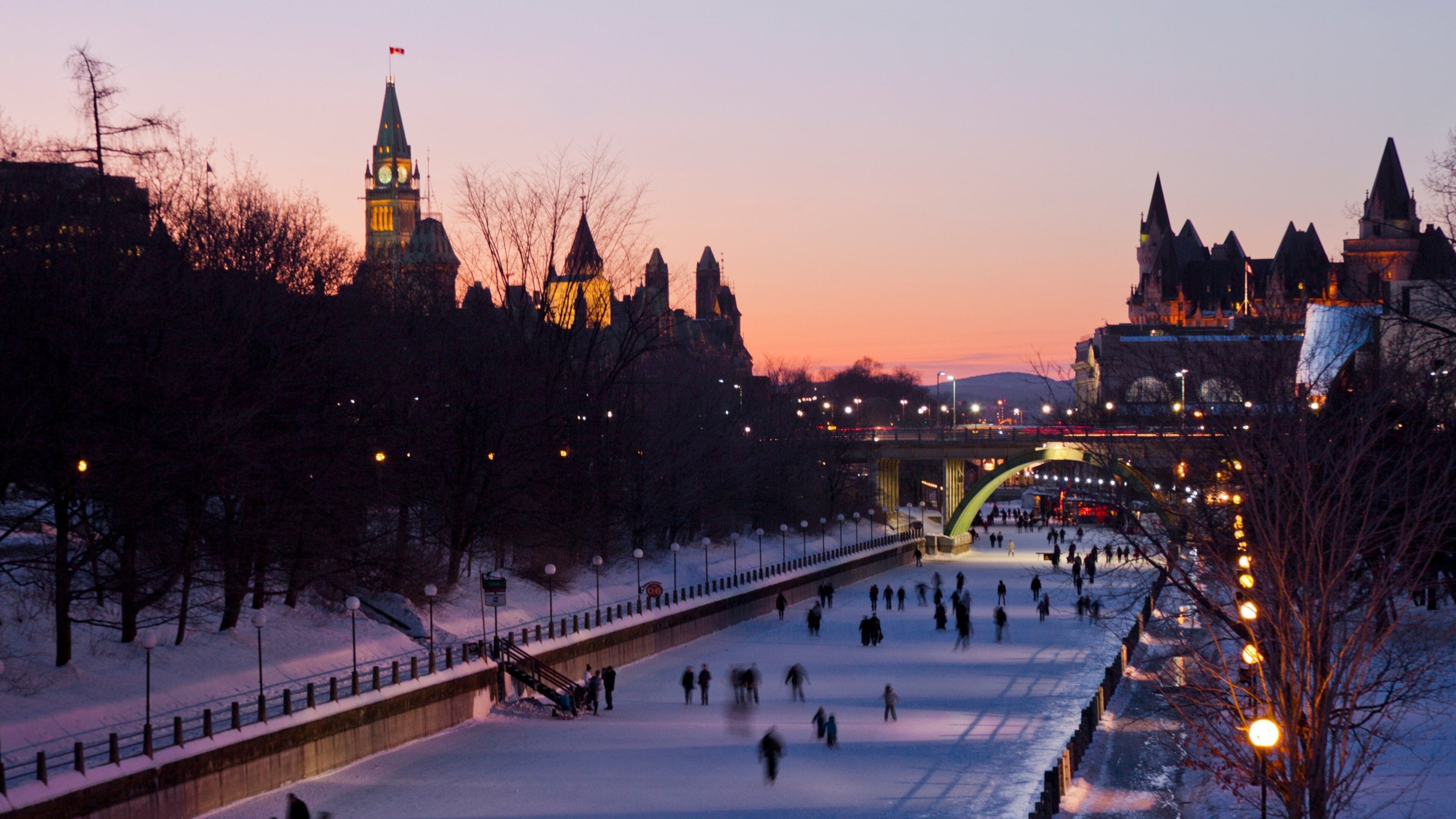 A snow-covered river with people ice skating, a bridge, and historic buildings in the background.