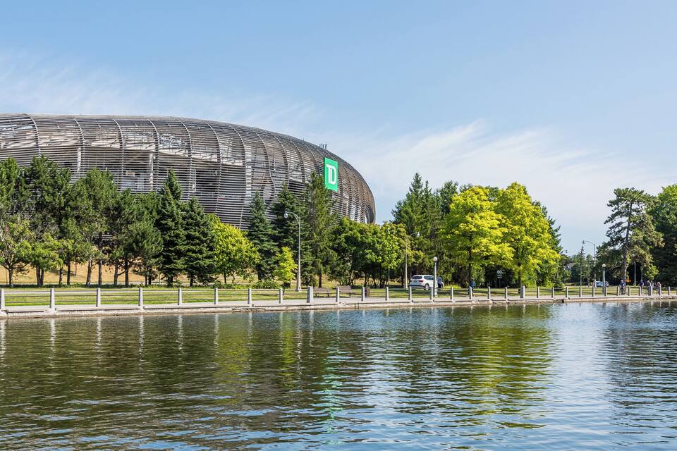 A modern stadium with a curved roof, surrounded by trees and a body of water.