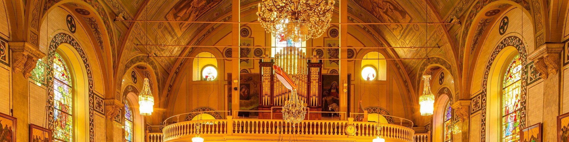 Interior imponente de igreja com tetos ricamente decorados, janelas de vitrais e um grande lustre.