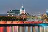 Un paysage urbain le long du littoral, avec un imposant bâtiment à coupole, un clocher d’église et un quai pour bateaux avec un toit rouge.
