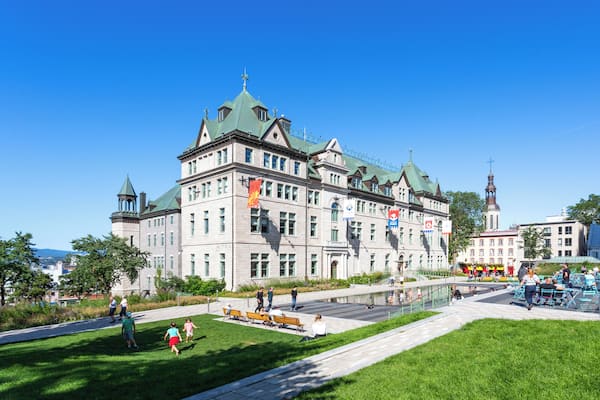 A historic building with green roofs and multiple flags, surrounded by a park with benches and people walking.