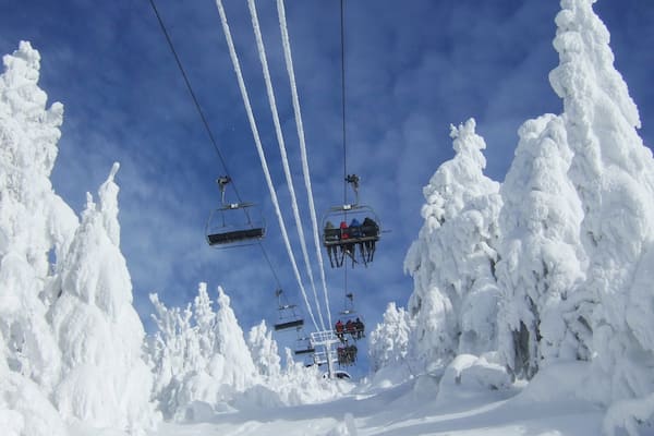 Eine Sesselbahn mit Gondeln, in denen Fahrgäste sitzen, umgeben von schneebedeckten Bäumen unter blauem Himmel.