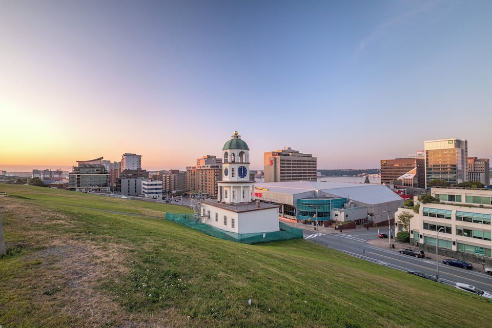 A cityscape with a prominent clock tower, modern buildings, and a green field in the foreground.