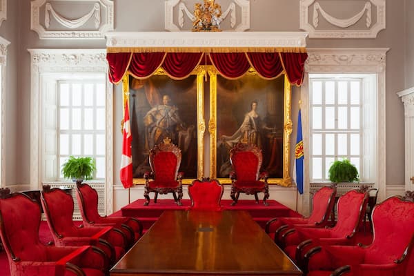 Une salle de réunion formelle, avec une longue table en bois, des chaises à revêtement rouge et un grand tableau représentant deux personnages accroché au mur.