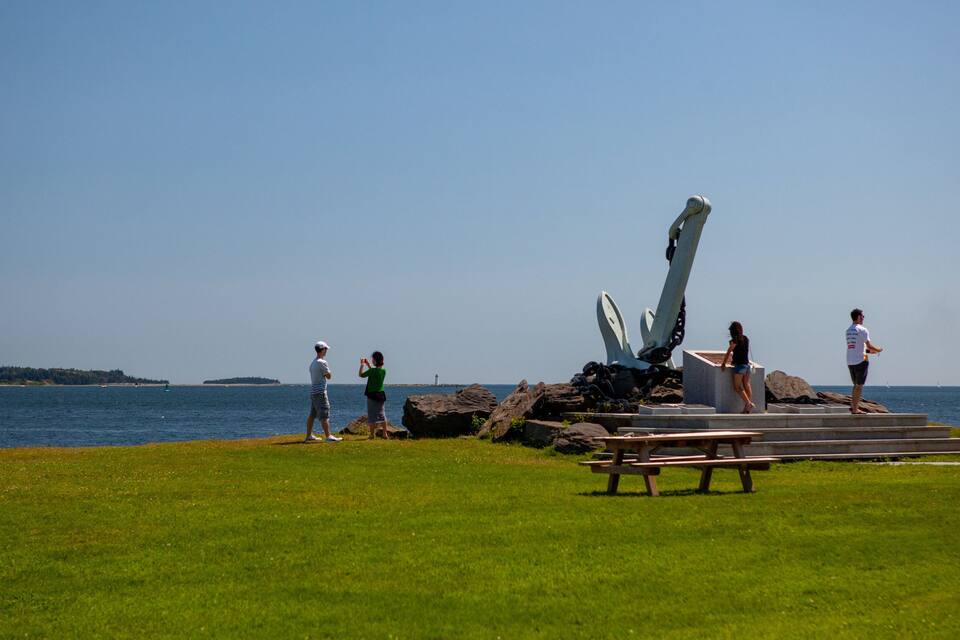 Un'area verdeggiante con un'ampia scultura a forma di ancora di nave, un tavolo da picnic e un faro in lontananza.