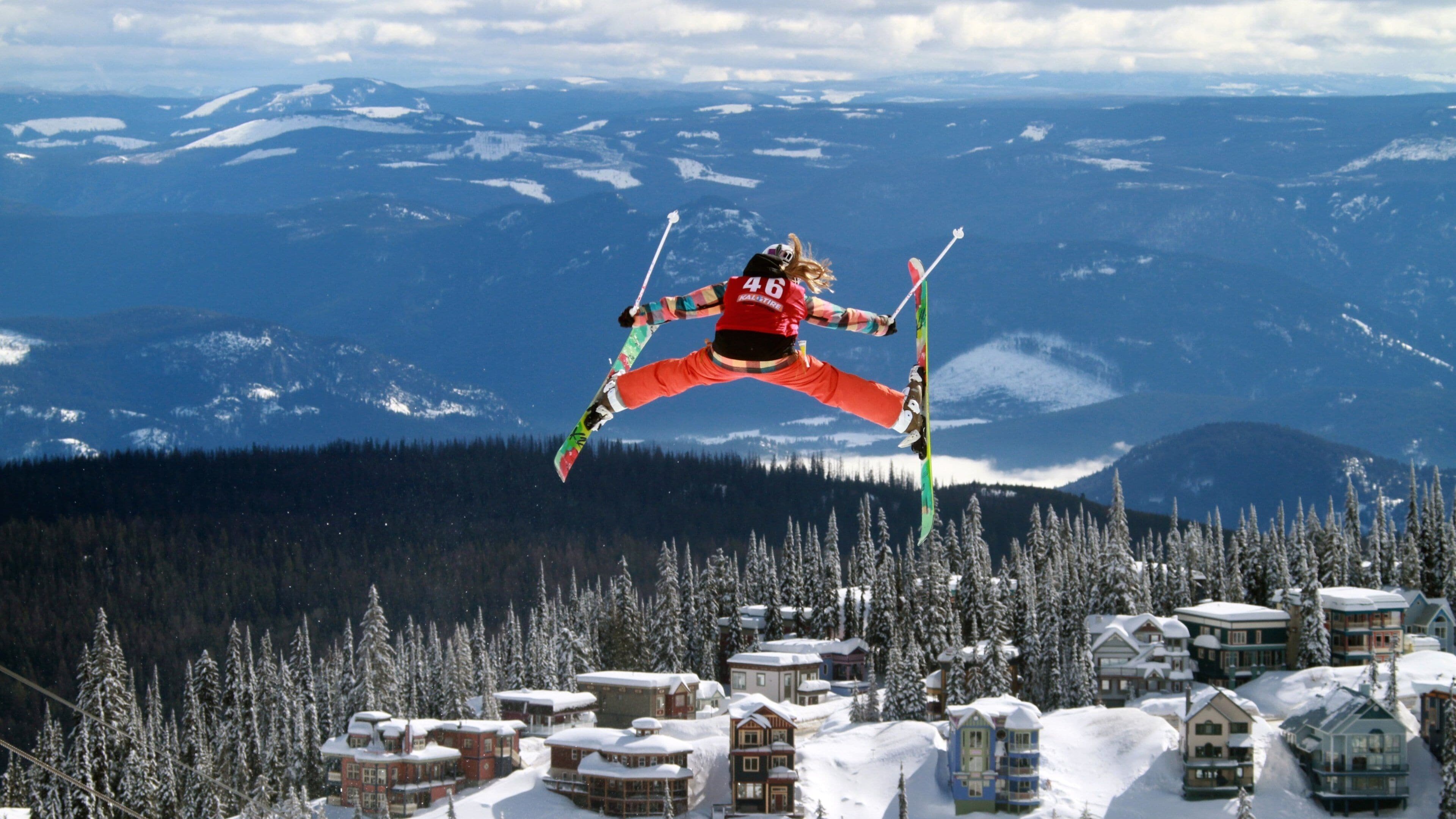 A skier mid-jump over a snowy village with mountains in the background.