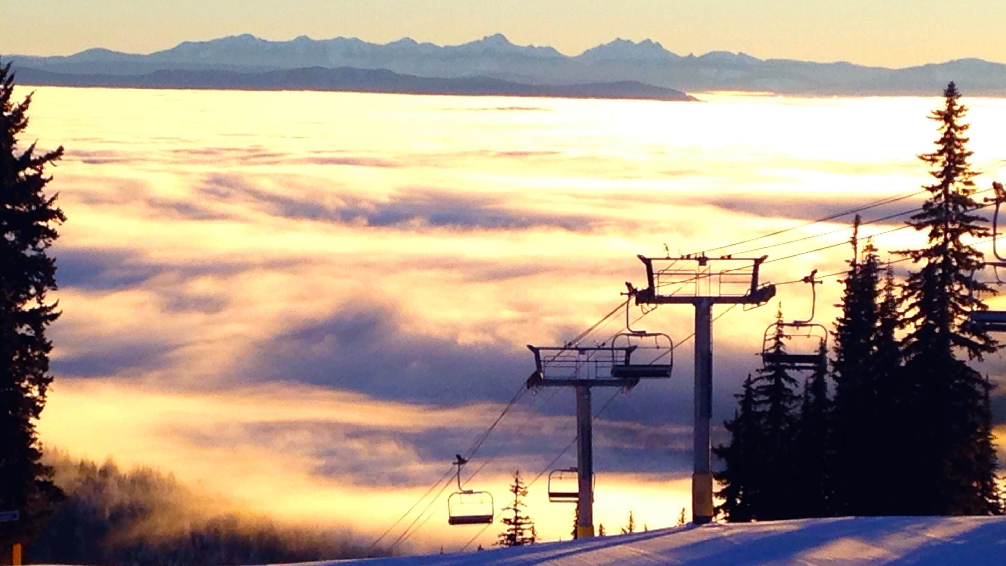 A ski lift with cables and chairs against a backdrop of mountains and a valley shrouded in clouds at sunrise or sunset.